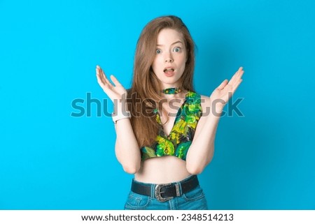 Surprised terrified young beautiful blonde woman wearing green top standing over blue studio background Gestures with uncertainty, stares at camera, puzzled as doesn't know answer on tricky question, 