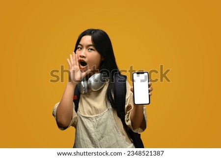 Cute Asian girl with backpack showing smartphone with blank screen on yellow background.