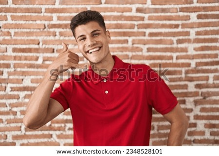 Young hispanic man standing over bricks wall smiling doing phone gesture with hand and fingers like talking on the telephone. communicating concepts.  Royalty-Free Stock Photo #2348528101