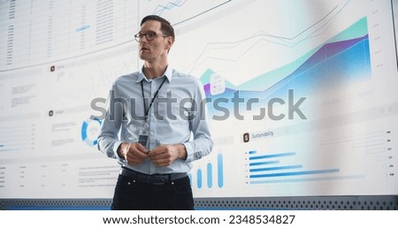 Male CEO Of Data Science Company Is Giving Presentation In Front Of Big Digital Screen With Graphs And Charts. Successful Caucasian Man Reviewing Business Accomplishments And Objectives In the Office.