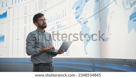 Indian Male Logistics Expert Holding Laptop Computer And Analyzing World Map On Big Digital Screen In Monitoring Office. Successful Man Developing New Efficient Routs For Global Product Distribution. Royalty-Free Stock Photo #2348534845