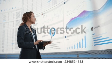 In the Monitoring Office: Successful Hispanic Businesswoman Holding Laptop Computer And Analyzing Data On Big Digital Screen With Graphs and Charts. Female IT Entrepreneur Running Software Company. Royalty-Free Stock Photo #2348534853