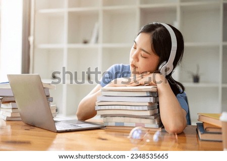 A beautiful Asian girl student is listening to music through her headphones, napping on stack of books, falling asleep while doing homework, and preparing for an exam in the library.