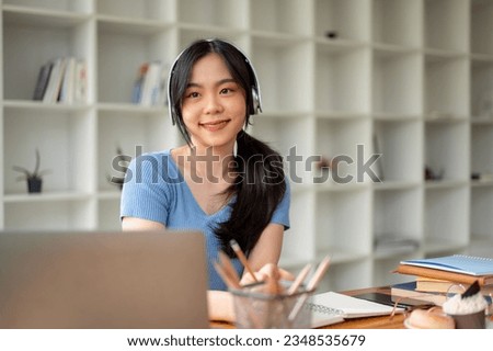 A beautiful young Asian female college student smiling, wearing headphones, while sitting at her study table in her room. Lifestyle concept