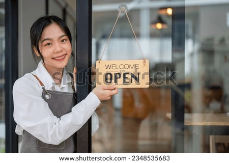 A portrait of a beautiful and friendly young Asian female restaurant staff or small business owner is hanging an open sign on the entrance door. welcome sign, open sign, available