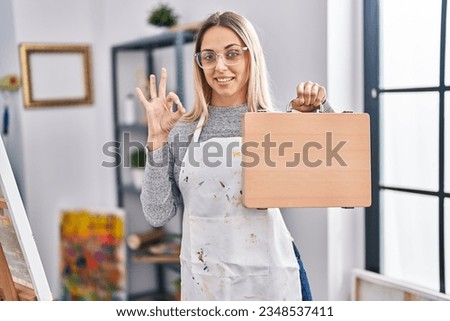 Young blonde painter woman holding wooden case at art studio doing ok sign with fingers, smiling friendly gesturing excellent symbol 