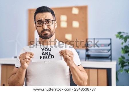 Young hispanic man with beard holding you are fired banner at the office skeptic and nervous, frowning upset because of problem. negative person. 