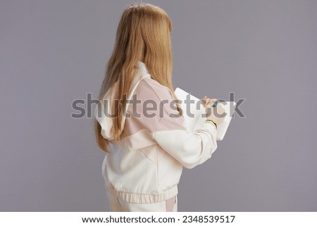 Seen from behind teen girl in beige tracksuit with workbooks writing against grey.