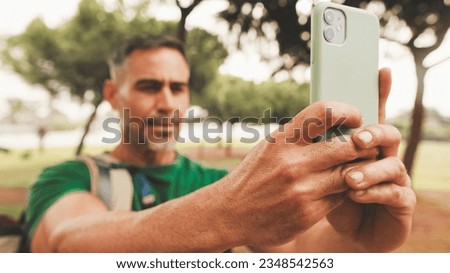 Middle-aged man dressed in casual clothes, with backpack on his shoulder, takes photo on mobile phone while standing in city park