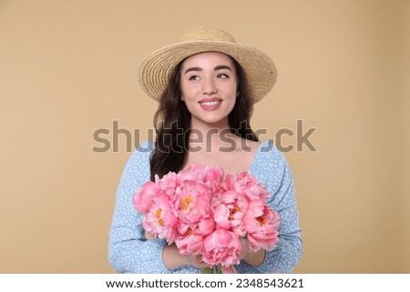 Beautiful young woman in straw hat with bouquet of pink peonies against beige background