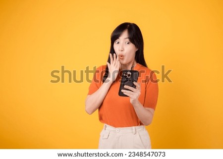 Excited Asian woman 30s, wearing orange shirt, using smartphone with fist up hand sign on vibrant yellow background. Thrilling new app experience. Royalty-Free Stock Photo #2348547073