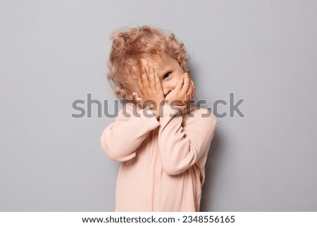 Adorable baby portrait. Shy baby girl with blonde wavy hair wearing rose shirt isolated over gray background hiding her face with arms looking through fingers to camera Royalty-Free Stock Photo #2348556165