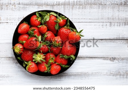Fresh organic strawberries in a black round plate on a light wooden background, top view, with space for text.
