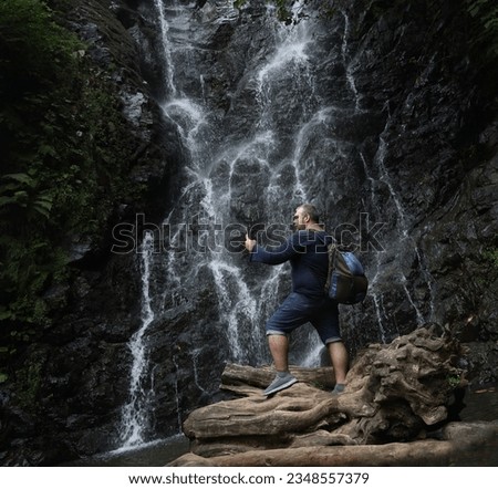 one, alone tourist man standing with backpack and looking at screen of  mobile phone. smart phone. front view of Mirveti waterfall. Georgia, Batumi.  Royalty-Free Stock Photo #2348557379