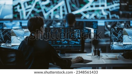 Back View Female Officer Using her Computer to Monitor Live CCTV Footage. Modern Monitoring Office With Big Digital Screen And Working Places of Employees With Desktop Computers and Displays Royalty-Free Stock Photo #2348561433