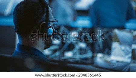 Technical Customer Support Specialist is Talking on a Headset while Working on a Computer in a Call Center Control Room Filled Display Screens and Data Servers. Footage from the Back Royalty-Free Stock Photo #2348561445