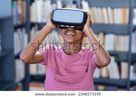 Smiling african american schoolboy using vr headset in school library, with copy space. Education, childhood, elementary school, communication, technology, inclusivity and learning concept.