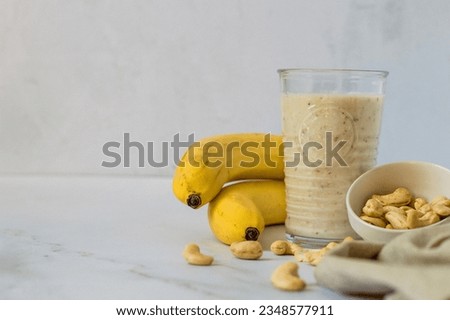 A glass of fruit smoothie from and a banana, a bowl of cashew nuts, on the table, the cooking process, on a light background
