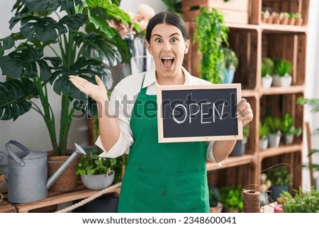 Young hispanic woman working at florist holding open sign celebrating achievement with happy smile and winner expression with raised hand 