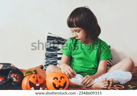 Child looks at the decorative spider on his shoulder. The girl is sitting on the sofa with pumpkin baskets for sweets, carnival glasses. Preparation for the Halloween holiday.