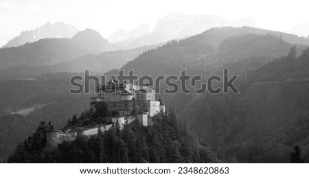 Rainy day panorama in austrian alps. Castle “Hohenwerfen“ in Werfen with silhouettes of mountain ranges in misty background. Black and white scenery with mystic atmosphere.