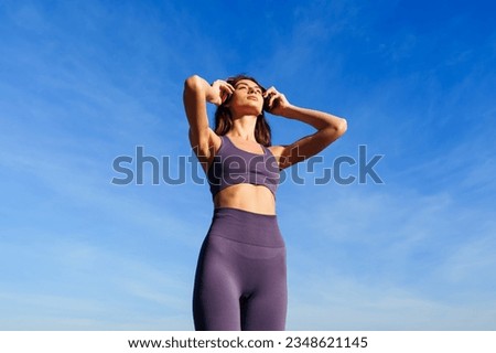 A young woman puts on headphones before training outside against a blue sky background.