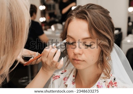 Shooting in a beauty salon. makeup artist applies makeup to a young beautiful girl bride. Royalty-Free Stock Photo #2348633275