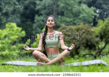 Beautiful young Indian woman doing yoga in the park. Sitting on a mat with closed eyes in the lotus position and meditating.