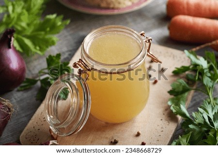 Bone broth in a transparent glass jar on a wooden table, with fresh vegetables
