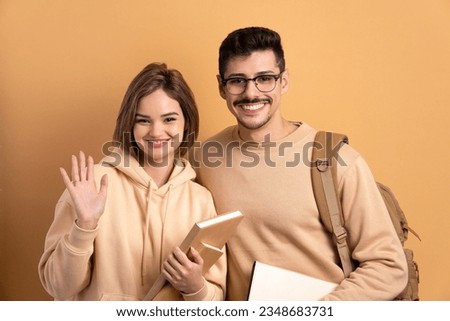 joyful student couple waving hello goodbye in beige studio background. learn, school, student life concept.