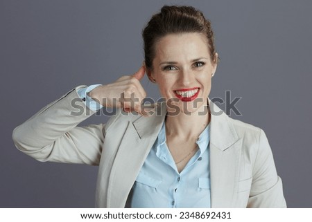 Portrait of smiling stylish female employee in a light business suit showing call me gesture against gray background. Royalty-Free Stock Photo #2348692431