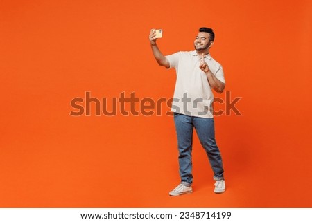 Full body young Indian man wears white t-shirt casual clothes doing selfie shot on mobile cell phone post photo on social network show v-sign isolated on plain orange red background. Lifestyle concept