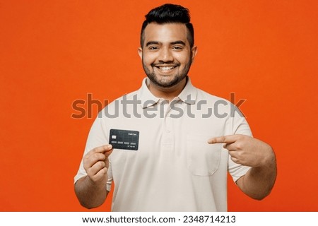 Young happy Indian man he wearing white t-shirt casual clothes hold in hand point on mock up of credit bank card look camera isolated on plain orange red background studio portrait. Lifestyle concept Royalty-Free Stock Photo #2348714213
