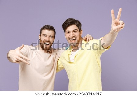 Two young smiling cheerful happy men friends together wearing basic casual t-shirt show victory v-sign gesture isolated on plain pastel purple color background studio portrait People lifestyle concept