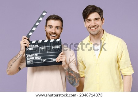 Young smiling happy men friends together wearing casual t-shirt tattoo translate fun holding classic black film making clapperboard isolated on purple background People lifestyle friendship concept.