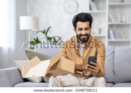 Joyful man at home on the sofa received a package, Latin American is satisfied with the purchase in the online store, writes a positive review using the application on the phone. Royalty-Free Stock Photo #2348738111