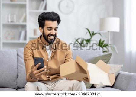Joyful man at home on the sofa received a package, Latin American is satisfied with the purchase in the online store, writes a positive review using the application on the phone.