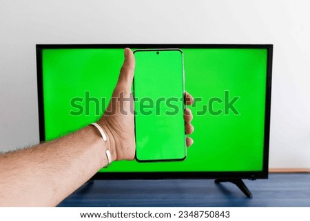 Hand holding a mobile phone with green screen with a green screen television in the background
