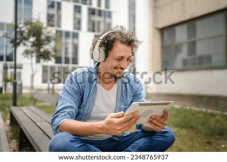 One caucasian man with curly hair outdoor in front of modern building use digital tablet and headphones to watch movie or series online stream or to have video call copy space