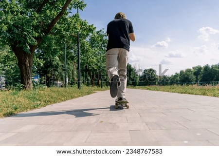 young guy skater rides skateboard on the path of the city park against the background of trees and sky