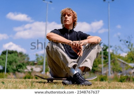 young serious guy skater with long hair sits on skateboard against the background of grass and sky