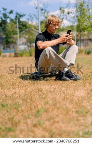 A young serious skater guy with long hair sits on a skateboard looking at his cell phone against background of grass and sky