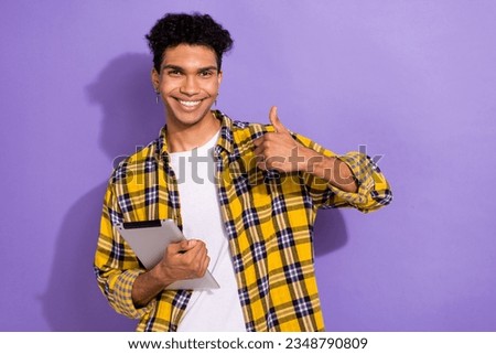 Photo of good mood cool guy dressed checkered yellow shirt reading modern gadget thumb up empty space isolated purple color background