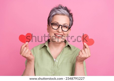 Photo of cute grandmother wear stylish khaki shirt holding two paper red postcards heart valentine day isolated on pink color background