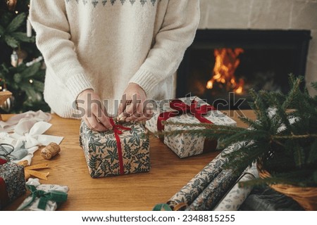 Woman wrapping stylish christmas gift with red ribbon on wooden table with festive decorations against fireplace in decorated scandinavian room. Merry Christmas! Hand holding present Royalty-Free Stock Photo #2348815255