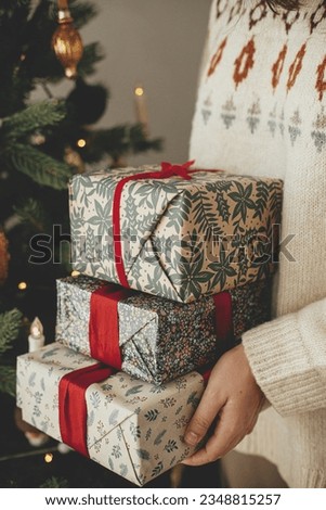 Woman in sweater holding stylish christmas gifts with red ribbon at modern decorated vintage tree in scandinavian room. Merry Christmas and Happy holidays!