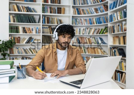 Serious concentrated man in headphones studying in library, hispanic student thinking and recording video course while sitting in university campus library.