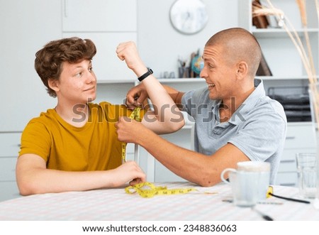 Father using measure tape for measuring girth of his son's biceps Royalty-Free Stock Photo #2348836063