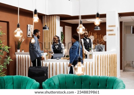 Young African American couple standing at hotel front desk with luggage, paying for room with credit card while checking out. Friendly smiling female receptionist serving tourists checking guests in Royalty-Free Stock Photo #2348839861