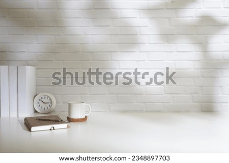 Minimal workplace, houseplant, notebook, glasses and coffee cup on white table with sunlight through blinds by the window.